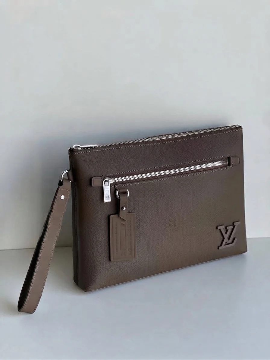 Brown grained leather clutch with a wrist strap and zip, standing against a pale wall